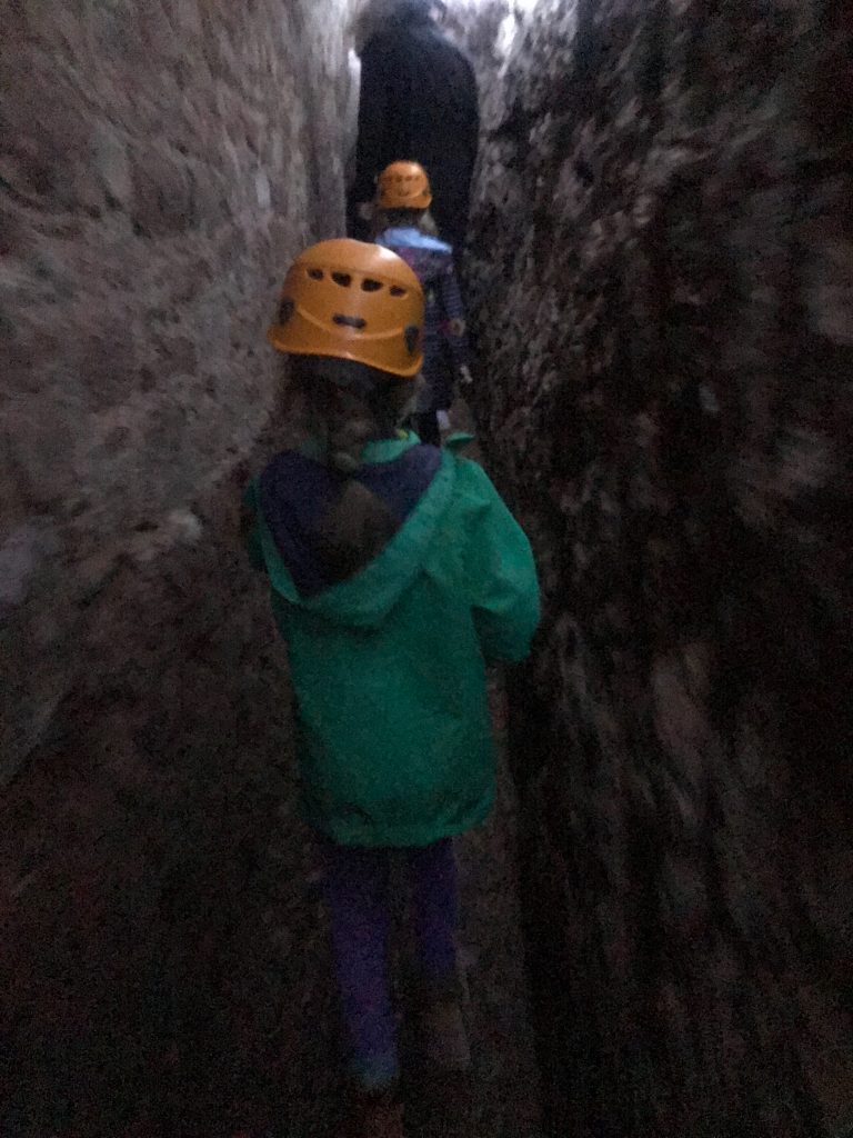 A slightly blurred photo taken in Exeter's Underground Passages showing the back of a small girl wearing a green coat and orange hard hat walking through a stone tunnel. In front of her is someone in a blue coat and orange hat.