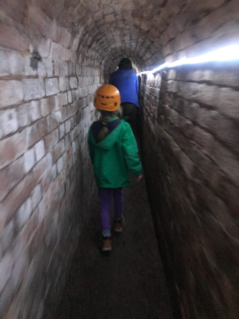 The same young girl in green coat and orange hat walks through a brick section of the tunnel. On the righthand side you can see a light rope along the tunnel wall. The photo is slightly blurred, but you can make out the adult in the tunnel infant of the girl who is having to stop slightly due to the low roof.