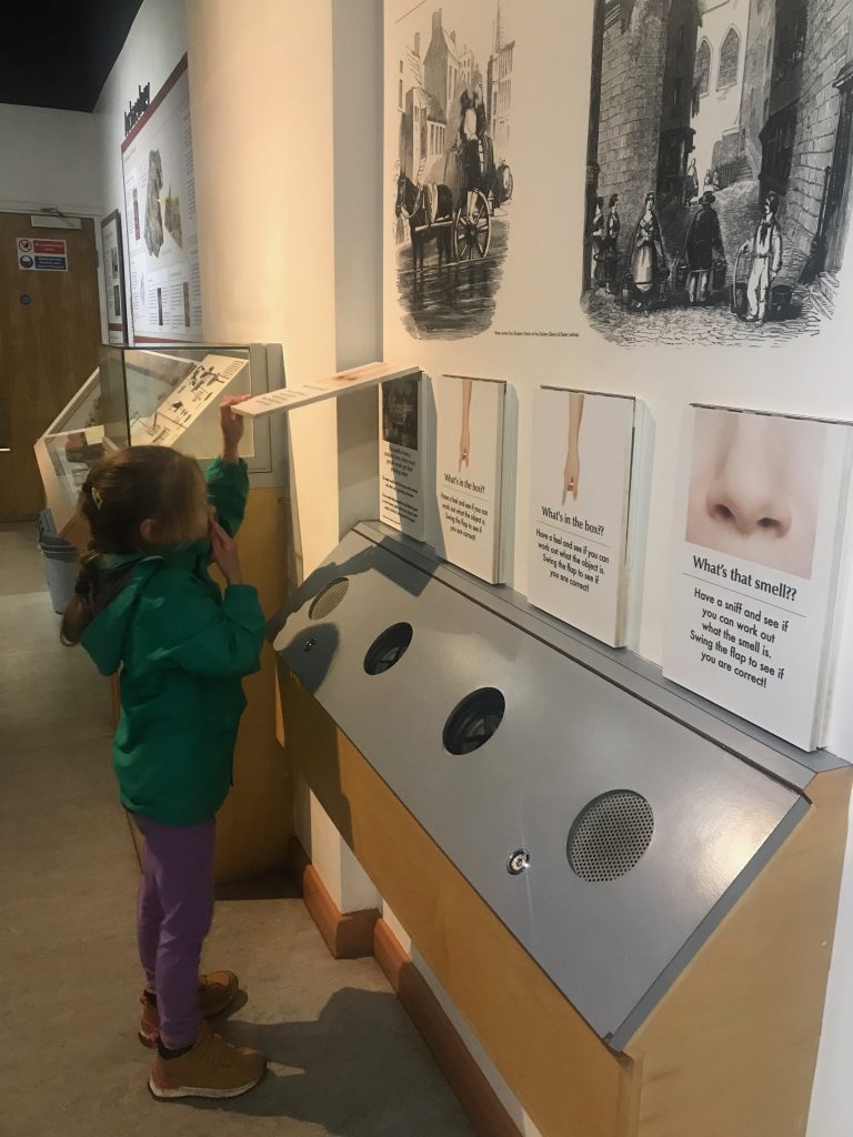 A five year old girl wearing a green coat holds her nose whilst lifting up a large flat to reveal what the smell is in the Exeter Underground Passages exhibition.